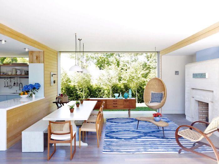 kuchyňa otvorená do obývačky, polootvorený model kuchyne, drevená barová stena, biely stôl a lavica, drevené stoličky, biely krb, ratanová hojdačka, veľké okno s výhľadom na zelenú záhradu