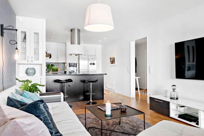 layout för litet kök öppen för vardagsrum, vit köksfront med svart centralö, ljus parkett, vit soffa, svart soffbord och grå matta, färgade kuddar