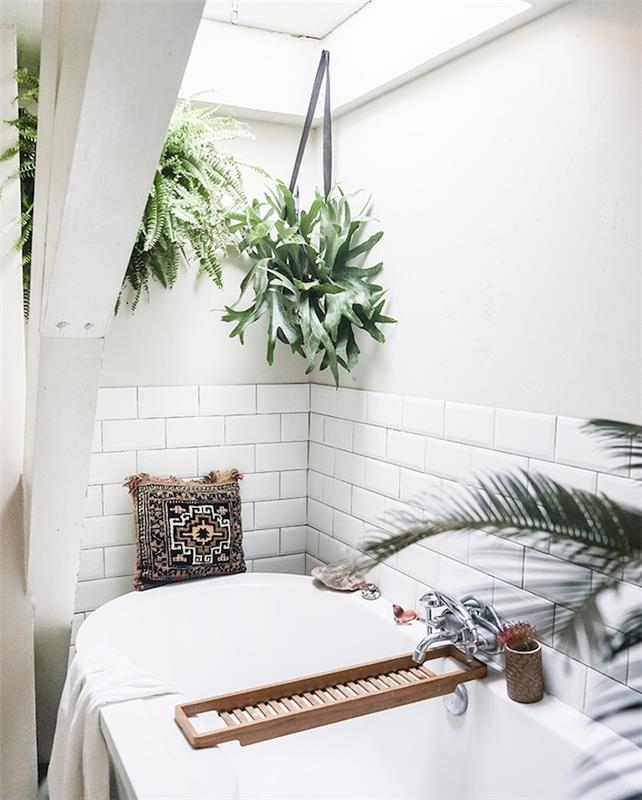 Zenová kúpeľňa v duchu dekorácie džungle so závesnými zelenými rastlinami v kvetináči, bielou vaňou, bielou dlaždicovou základňou, orientálnym dekoratívnym vankúšom