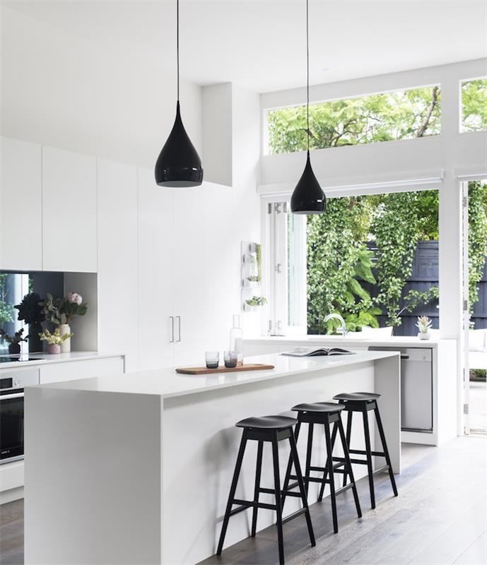 modernt vitt kök med vit centralö omgiven av svarta pallar, mörkt trägolv, vitt köksskåp, svarta hängande lampor
