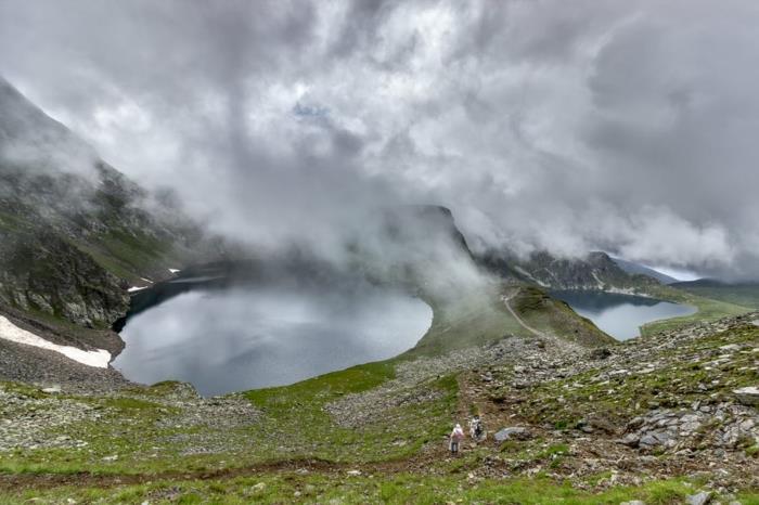 dimmig sjö i berget, grönt gräs, grå stenar, vackert landskap, utsikt från ovan, pärlgrå himmel, upphetsade turister klättrar till toppen