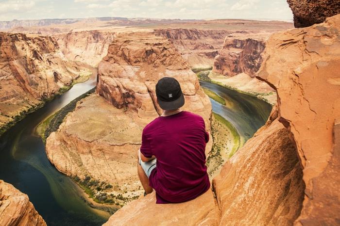 paradisiskt landskap, man i en vinröd T-shirt sitter på kanten av en klippa ovanför en kanjon, vatten med gröna och blå nyanser