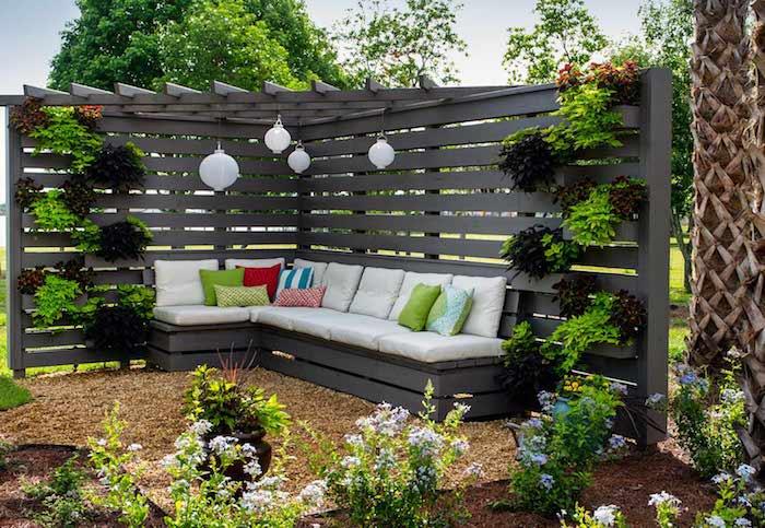 moderná pergola s vertikálnou stenou rastlín a sivou sedačkou s bielym vankúšom na sedenie a farebnými dekoratívnymi vankúšmi, záhradné rastliny