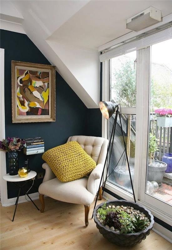 liten flytande bokhyllvägg med benvit fåtölj och gråblå vägg med färgglad konstram och stor inomhus trädgårdskruka