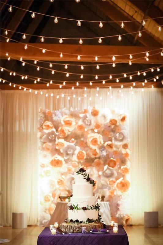 stena na svadobnom pozadí zdobená papierovými kvetmi, svetelné girlandy na ozdobu stropu, svadobná torta na drevenom poli