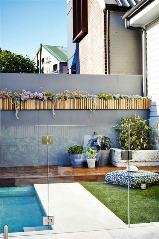 vonkajšia dekorácia steny, vonkajšia obklad stien s držiakmi rastlín z ľahkého dreva, záhradný plot s bazénom v pastelovo šedej farbe, zelená tráva s veľkým štvorcovým vankúšom na relaxáciu