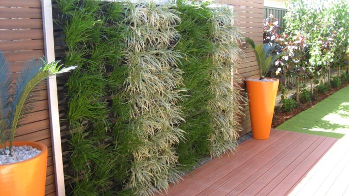 vertikálna záhrada, stĺpové panely so zelenými rastlinami, dva kvetináče v kónickom tvare v oranžovej farbe s palmami, zelená stena