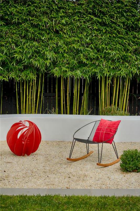 priečka na rastliny s bambusovými stonkami, uvoľnený záhradný kútik s kreslom z čierneho kovu a svetlého dreva, červená látková guľa, dekorácia dizajnu, jemný piesok, udržiavaná zelená tráva, vonkajšia stena rastliny