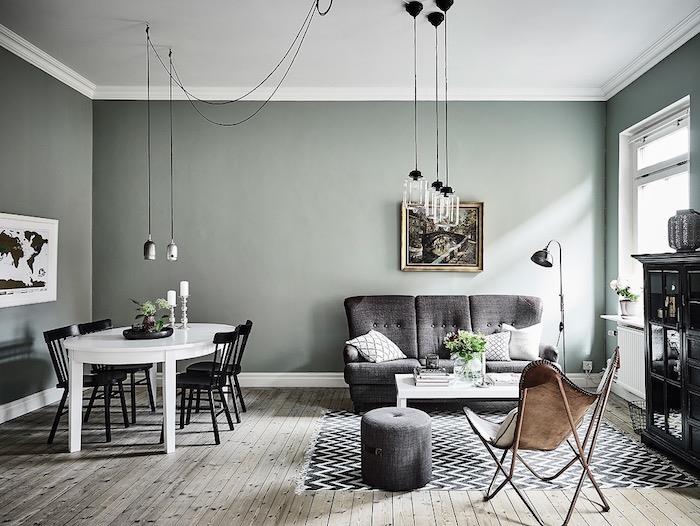 celadon verdigris vymaľuje steny v škandinávskej obývačke s jedálňou, parkety zo svetlého dreva, originálne závesné svetlá, sivú pohovku a taburetku, čierno-biely koberec v tvare šípky, koženú stoličku, biely stôl a čierne stoličky