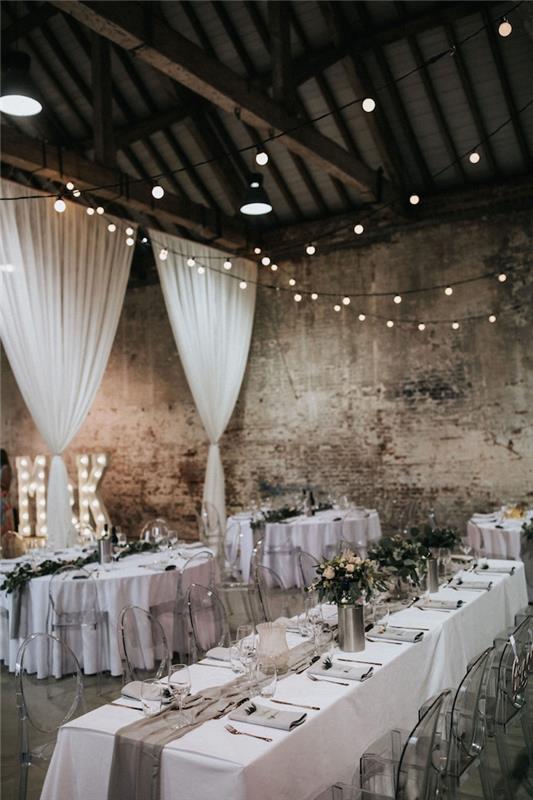 industriálna svadba stará továreň, vyzdobená bielymi priehľadnými závesmi, stoly ozdobené bielymi obrusmi a sivou látkou, kovová váza, interiérová dekorácia zo svetlej girlandy