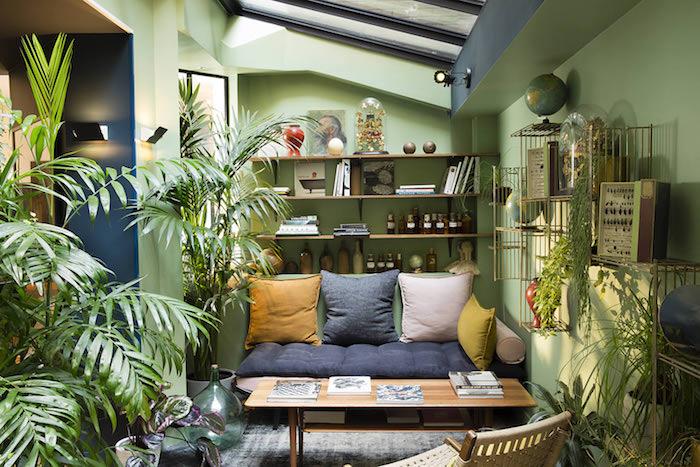 zelená stena, drevený stôl a sivá a zelená pohovka, sivý, biely a žltý vankúš, množstvo rastlín v kvetináčoch na podlahe, tropický dekor