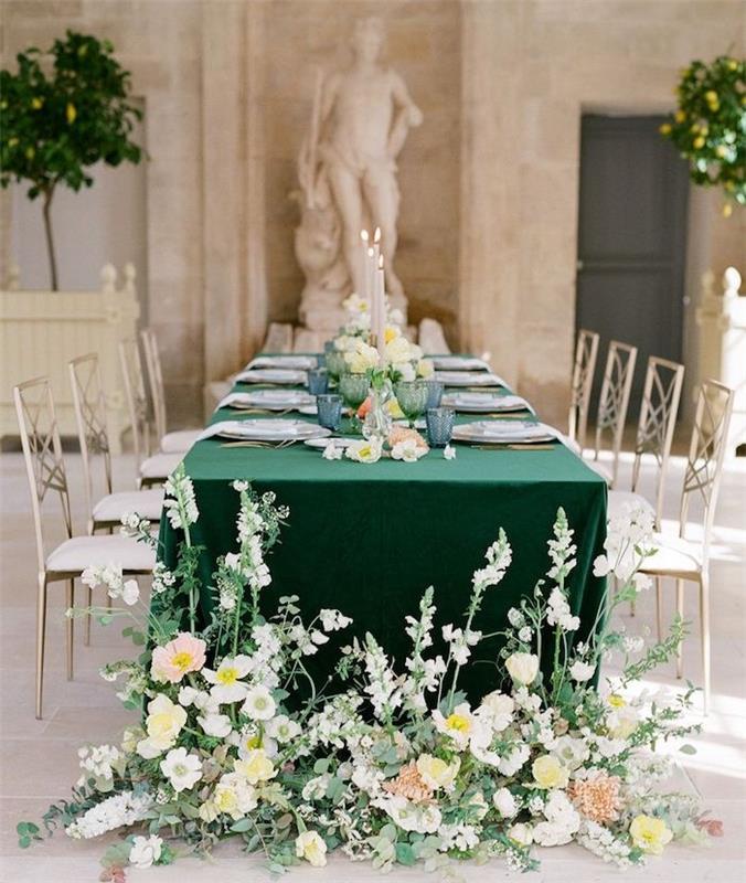 modré okuliare a biele taniere na zelenom obruse, stôl obklopený striebornými a bielymi stoličkami, kvetované nohy stola, rímske sošky