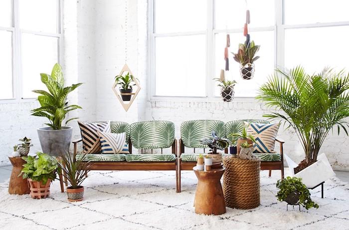 tropická výzdoba obývačky s drevenou pohovkou, operadlom a vankúšmi s palmou, niekoľkými závesnými a umiestnenými kvetináčmi, bielou tehlovou stenou a bielym kobercom