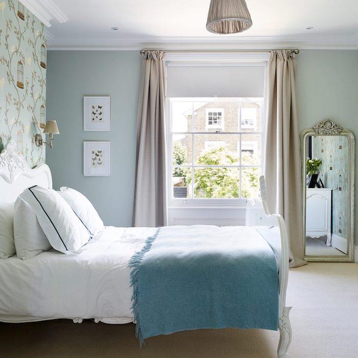 kačacia modrá tapeta do spálne, vzor chinoiserie, vtáčia klietka a kvitnúce konáre, biela posteľná bielizeň s modrou prikrývkou, baroková posteľ a zrkadlo