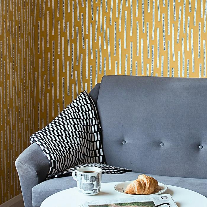 fiberduk tapeter i gult och vitt, med humoristiska budskap, pastellblå sammetsoffa, litet runt och vitt bord med croissant och kaffe