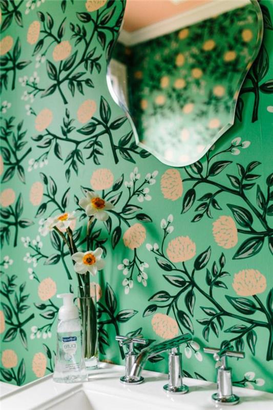 Liberty tapet i reseda och beige, med ramlös spegel i barockform, vintage vit handfat, transparent glasvas med tre blommor, nyklassisk atmosfär