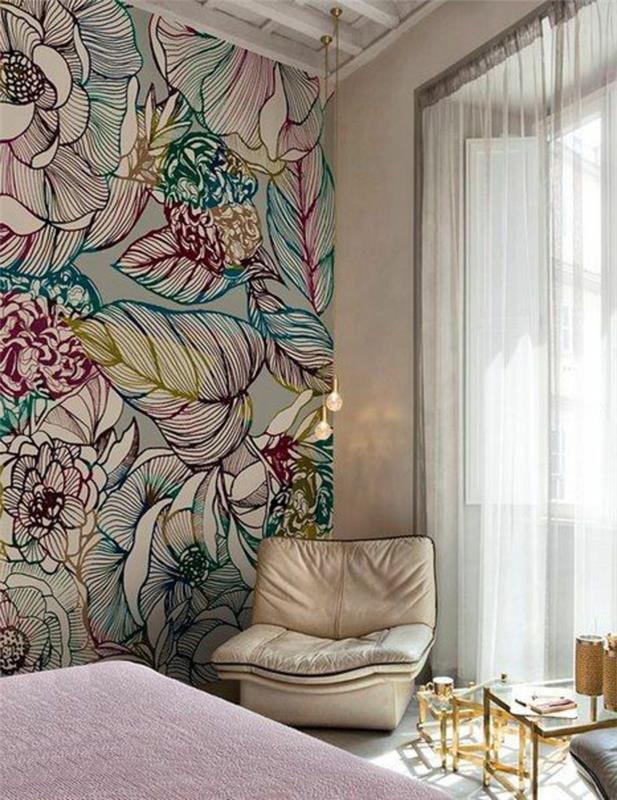 trompe l'oeil tapet i ett sovrum för vuxna, stora blommor och löv i ljusa färger, stor fåtölj i konstläder i beige färg, litet soffbord i vitt glas och ben av bronsmetall, tak med vita balkar och hängande lampor glödlampformade