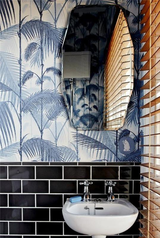 Liberty tapet med stora palmblad i blå färg, spegel i geometrisk form, hälften av väggen i svart tegel, liten vit diskbänk med två kranar, persienner i beige