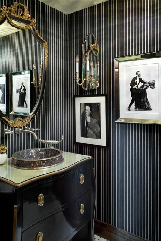 trompe l'oeil tapet, vägg i fina svartvita ränder, klassisk möbel i svart med glansig blank yta, renässansspegel, med gyllene dekorelement, runt format handfat, med mosaikbitar, vägglampa av spegeltyp med spegel för reflekterande ljus , två tavlor i retrostil i svartvitt