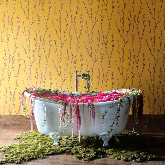 liberty tapet i senapsgula och små känsliga blommor i rosa, badrum med vitt ovalt badkar i vintage-stil, golv täckt med mörkbrun PVC-parkett, dekorationsörter och konstgjorda blommor i grönt och rosa runt badkaret