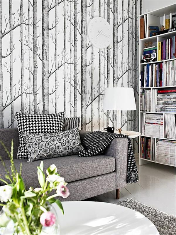 fiberduk tapeter med träd i en skog, i grå och svarta färger, grå soffa med stora dynor med fyrkantiga och geometriska mönster, låg golvlampa med trumskärm, placerad på ett runt vitt bord, grå matta