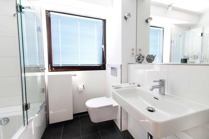 layout av ett litet vitt badrum med stor spegel, badrumsutrustning med badkar, dusch och upphängd toalettskål