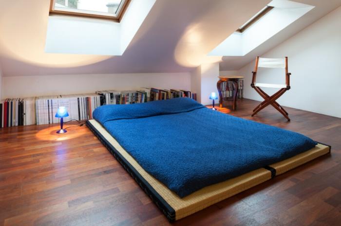 säng på golvet, brun träparkett, blå sänglinne, förvaring för böcker på golvet, fällstol, vit väggfärg, sovrumsdekoration under sluttningen