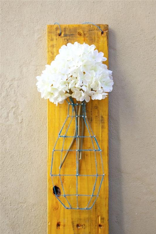 vacker dekorativ vas i blå tråd och spikar fyllda med vita blommor på en träbräda målad i gult