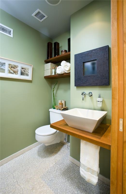 dekor toalety so zelenými stenami a bielymi dlaždicami, obmedzený priestor na skladovanie s poličkou z tmavého dreva