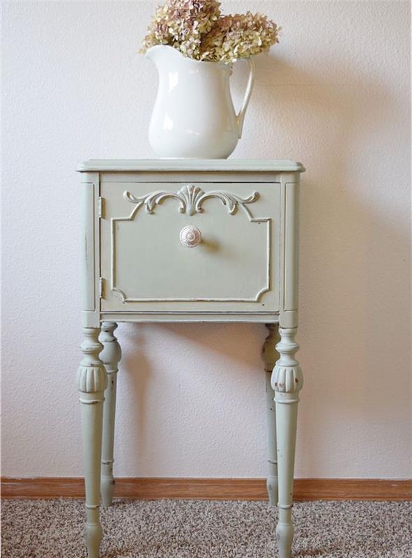 patina kus nábytku, malý príručný stolík so zásuvkou prelakovaný celadonskou zelenou farbou s bielym gombíkom, kytica kvetov v bielej váze, sivý koberec