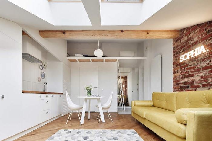 säng över toaletten i en 20 m2 studio med vitt kök, vitt matbord och stolar, synlig träbjälke, gul soffa, tegelvägg