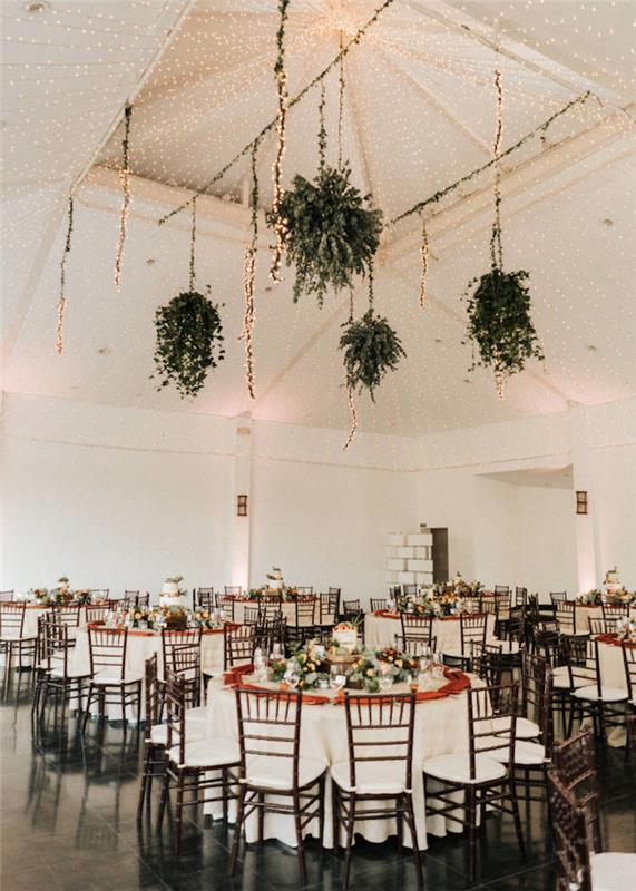 závesné kvetináče zo zelených rastlín a strop zdobený svetlými girlandami nad stolmi v bielych obrusoch a drevenými a bielymi stoličkami na čiernej dlažbe