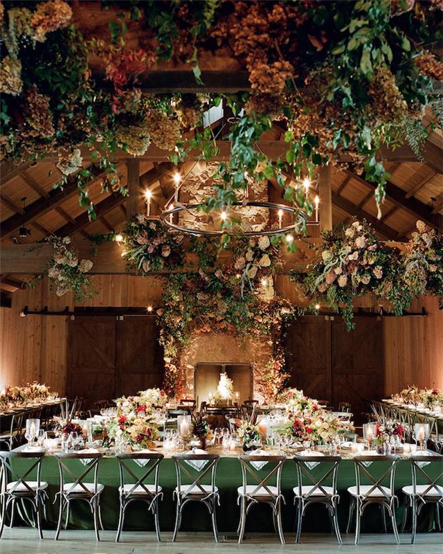 svadba v starých zalesnených stodolách s kvetinovou výzdobou stropu, svadobným krbom a stolmi v kvetovanej zelenej