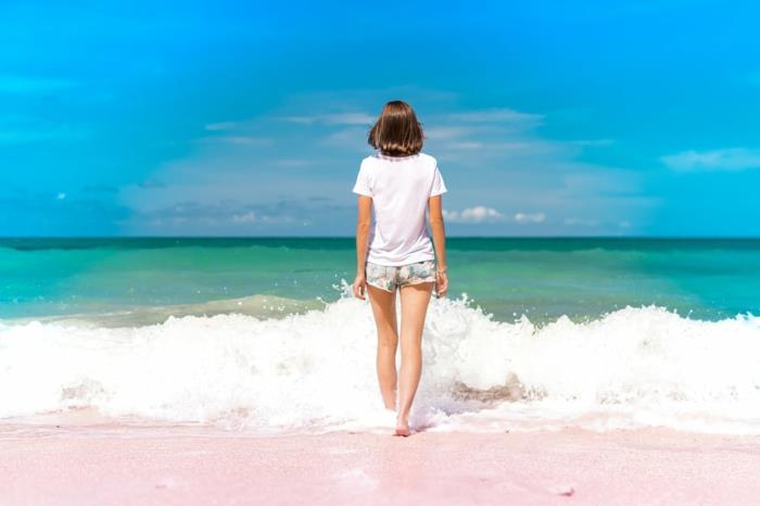 en liten flicka som kommer in i havet, lyfter foten på den skummiga vågen som anländer till stranden, horisonten i blått och grönt, vita moln, beige sand