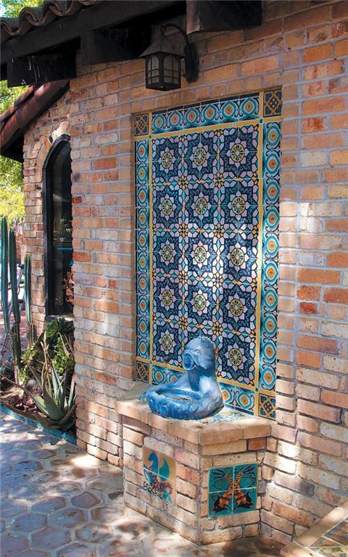 Ako obliecť vonkajšiu mozaikovú stenu, prvky mozaiky v modrej, žltej a oranžovej farbe, kompozícia vložená do červenej tehlovej steny, svetlo lampáša, tyrkysovo modrá keramická soška