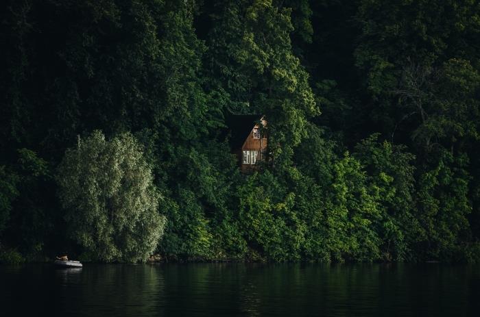 dvojpodlažný drevený dom postavený v lese nad jazerom, čln vo vode odrážajúci zelené stromy