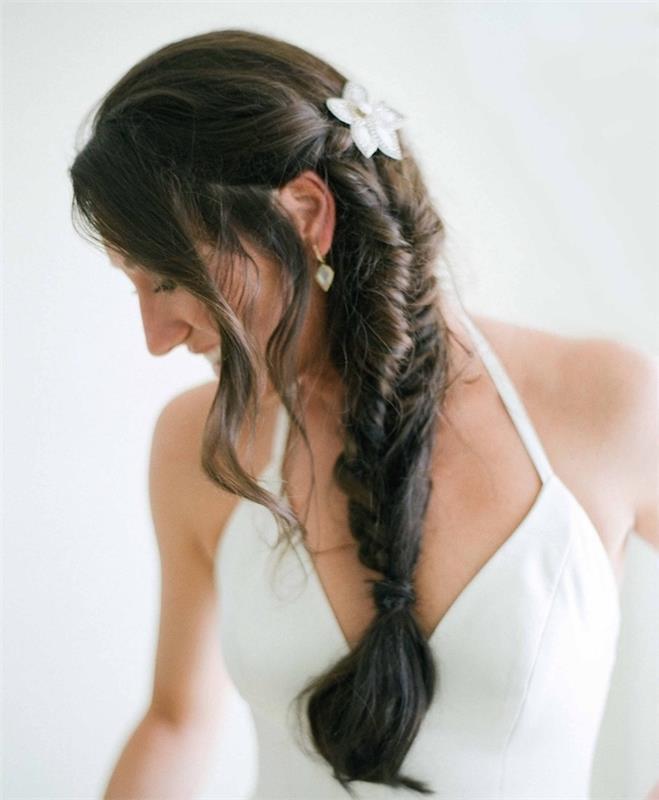 brudfläta på mörkt brunt hår, lösa trådar och en vit blomsterdekoration, enkel brudklänning