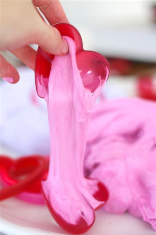 كيفية صنع السلايم بكريم الحلاقة ، تمسك باليد بنصف وعاء بلاستيكي أحمر ، على شكل قلب ، ينزف سلايم وردي رقيق