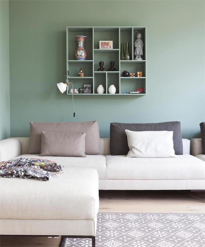 Zelená dispozícia obývačky Celadon, sivobiela rohová sedačka, sivé vankúše, sivobiely koberec, svetlé parkety a zelená otvorená polica, úložný priestor pre budhistické dekoratívne doplnky