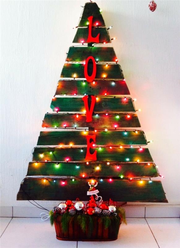 teplý vianočný stromček s farebnými žiarovkami, naukladané vianočné hračky a zeleň, veľké ozdobné písmena, paletový stromček