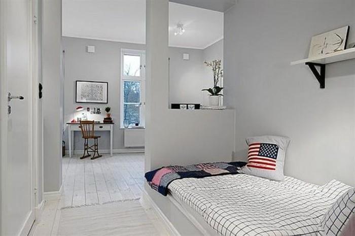 وسادة علم الولايات المتحدة ، على سرير بغطاء متقلب ، داخل غرفة بجدران رمادية شاحبة ، ورف كتب واحد ، أفكار لتجهيز الغرفة ، مكتب أبيض مع كرسي بني في الخلفية