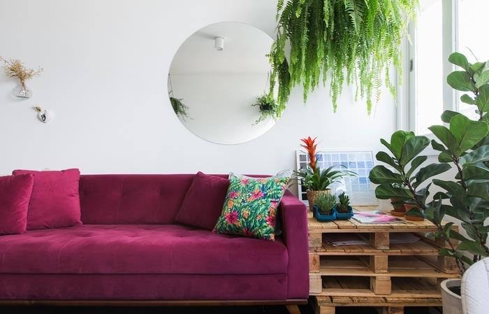 paletový stôl v surovom vzhľade umiestnený vedľa modernej fuchsiovej ružovej zamatovej sedačky pre príjemný kontrastný efekt v tejto tropickej obývačke