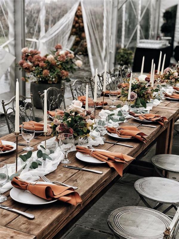 Téma svadobnej dekorácie stolu na jeseň s kvetinovými aranžmánmi z hnedých, bielych a ružových kvetov na stole zo surového dreva s bielym závojom