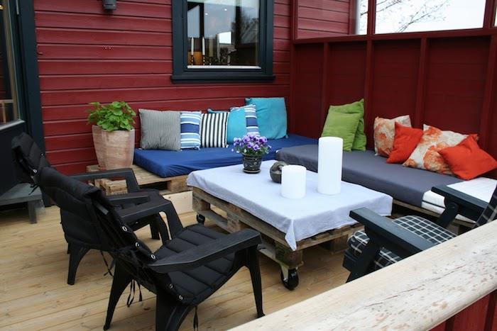 príklad krytej terasy s nízkym paletovým stolom a rohovou sedačkou v paletách, čiernych stoličiek, množstva farebných dekoratívnych vankúšov