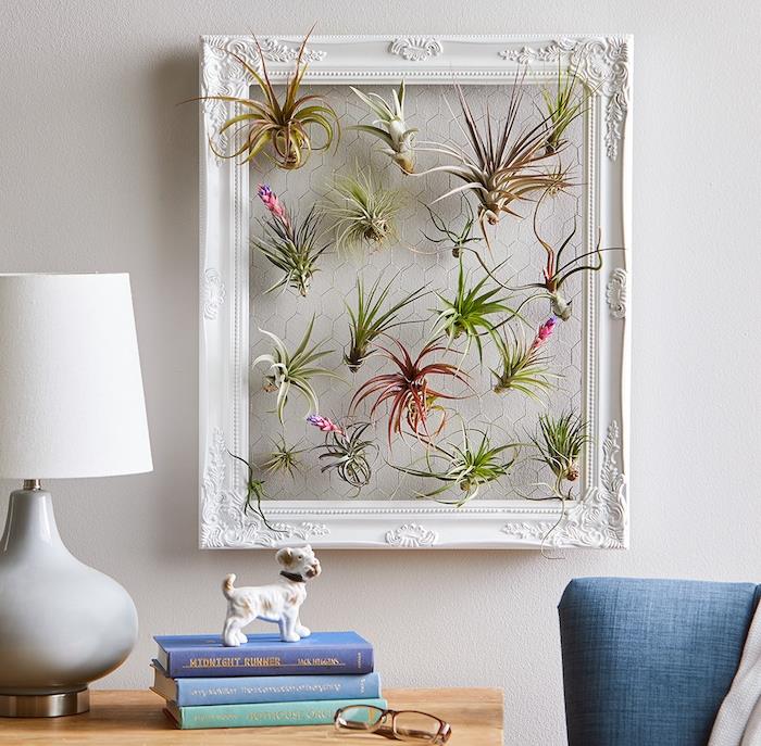 dekoratívna rastlinná maľba so vzduchovými kvetmi zavesená na plote zarámovanom na bielej stene, stoh kníh, figúrka psa, pôvodná izbová rastlina