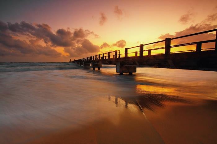 drömlandskap, bro i havet, orange och gul himmel, våg som lugnt omfamnar stranden, havslandskap, paradisiska öar