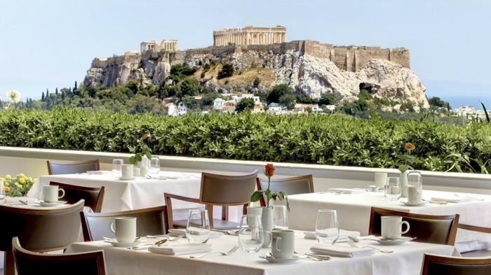 zelená stena ako zábradlie v reštaurácii v Aténach, svetlo hnedé stoličky, nádherný výhľad na Akropolu, bezoblačná modrá obloha