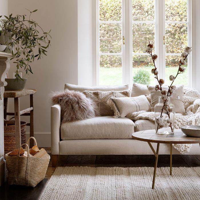 váza s bavlnenými ratolesťami na minimalistickom drevenom stole Pletený koberec sivobielej sedačky zdobený hrubým plédom a vankúšmi, elegantný dekor do obývačky