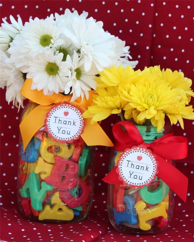 sklenená nádoba naplnená plastovými písmenami abecedy so stužkovou ozdobou a visačkou na darčeky, kyticou bielych a žltých kvetov